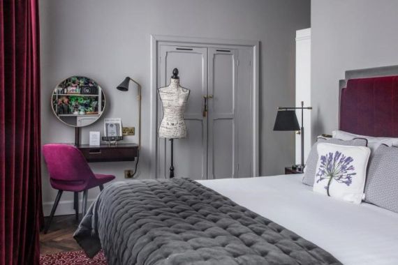 Deze stijlvolle Wilder Townhouse-slaapkamer heeft een pluchen bed met een grijze quilt en kussens met een plantenthema. Een paspop staat bij een kaptafel met een ronde spiegel en een paarse stoel, terwijl een zwarte lamp elegantie toevoegt. Donkerrode gordijnen bieden hotelachtige luxe tegen grijze muren en deur. | Echt Ierland Vakanties