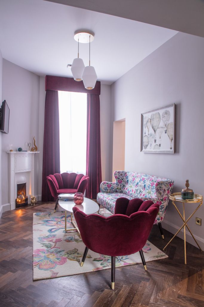 Een stijlvolle woonkamer in het Wilder Townhouse heeft twee kastanjebruine stoelen en een bloemenbank rond een ronde salontafel. Een kleurrijk kleed ligt op de visgraathouten vloer. Een verlichte open haard met decoratie staat in de hoek, terwijl kunstwerken van heteluchtballonnen de muur sieren naast grote ramen met bordeauxrode gordijnen. | Echt Ierland Vakanties