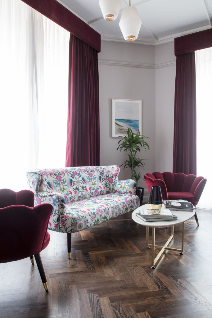 Ervaar de elegantie van een Wilder Townhouse-woonkamer, met een bank met bloemenmotief en twee bordeauxrode fluwelen stoelen. Een ronde salontafel met glazen blad bevat kaarsen en decoratie. Grote ramen met doorschijnende gordijnen complementeren de bordeauxrode draperieën, terwijl een potplant en een landschapsschilderij de lichtgrijze muren sieren. | Echt Ierland Vakanties