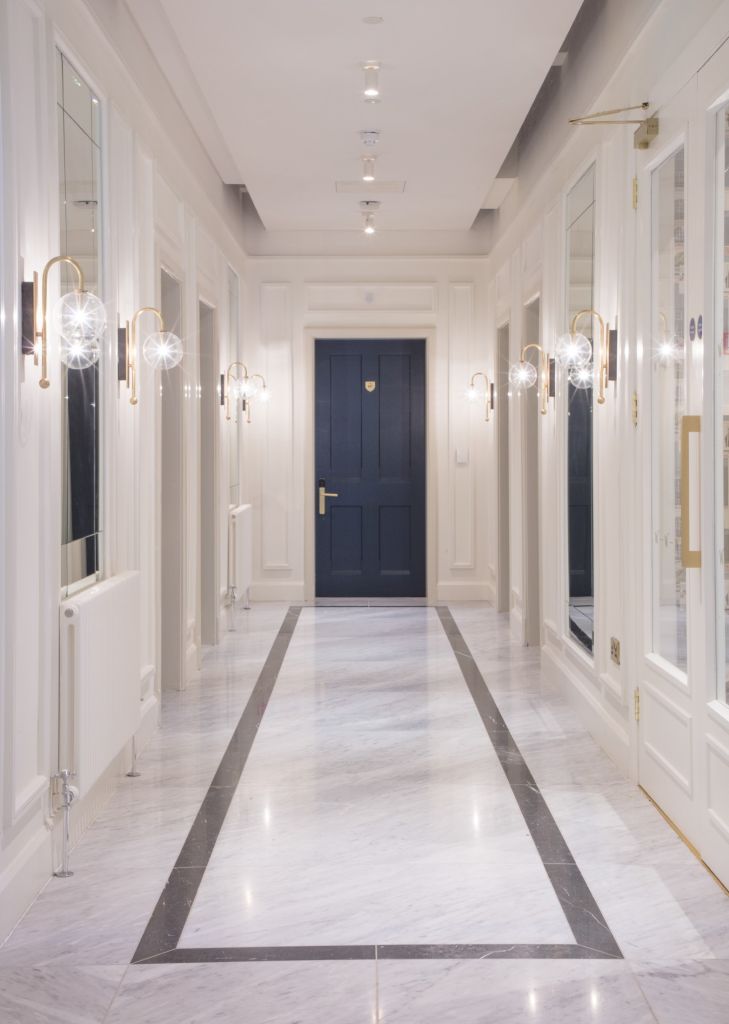 De elegante hal van het Wilder Townhouse heeft een witte marmeren vloer en zwarte afwerking die naar een gesloten zwarte deur leidt. Muren versierd met verticale spiegels en metalen wandlampen die zacht licht uitstralen. Een smalle radiator is zichtbaar aan de linkerkant onder inbouwverlichting, wat een lichte, luchtige sfeer biedt die past bij een hotel. | Echt Ierland Vakanties