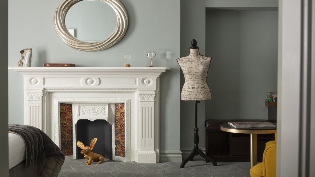 In het Wilder Townhouse is een stijlvolle kamer voorzien van een witte, sierlijke open haard met daarin een gouden konijnensculptuur. Boven de schouw hangt een ronde spiegel, versierd met een kleine wereldbol en een zandloper. Rechts staat een vintage paspop bij een gele fauteuil naast een kleine ronde tafel. | Echt Ierland Vakanties