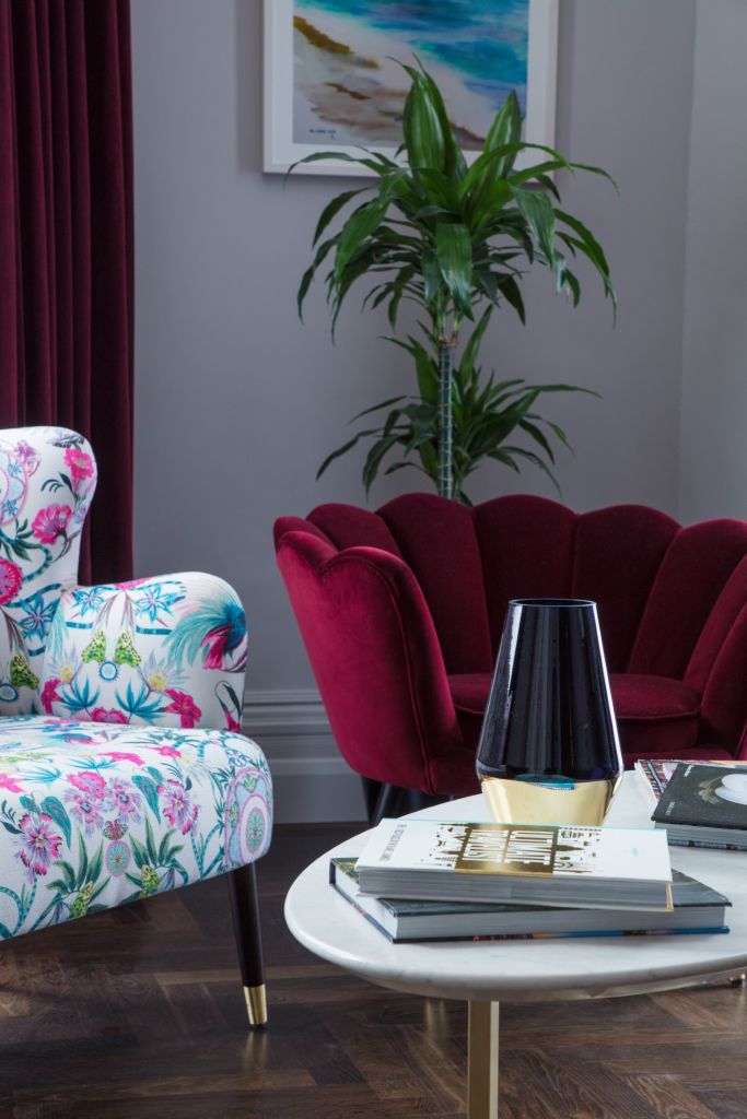 Een stijlvolle woonkamer in het Wilder Townhouse heeft een bloemenfauteuil en een dieprode fluwelen bank. Een hoge groene plant staat ertussen, terwijl een ronde salontafel van wit marmer een zwart-gouden lamp en wat boeken bevat. Een kleurrijk schilderij van de kust siert de grijze muur. | Echt Ierland Vakanties