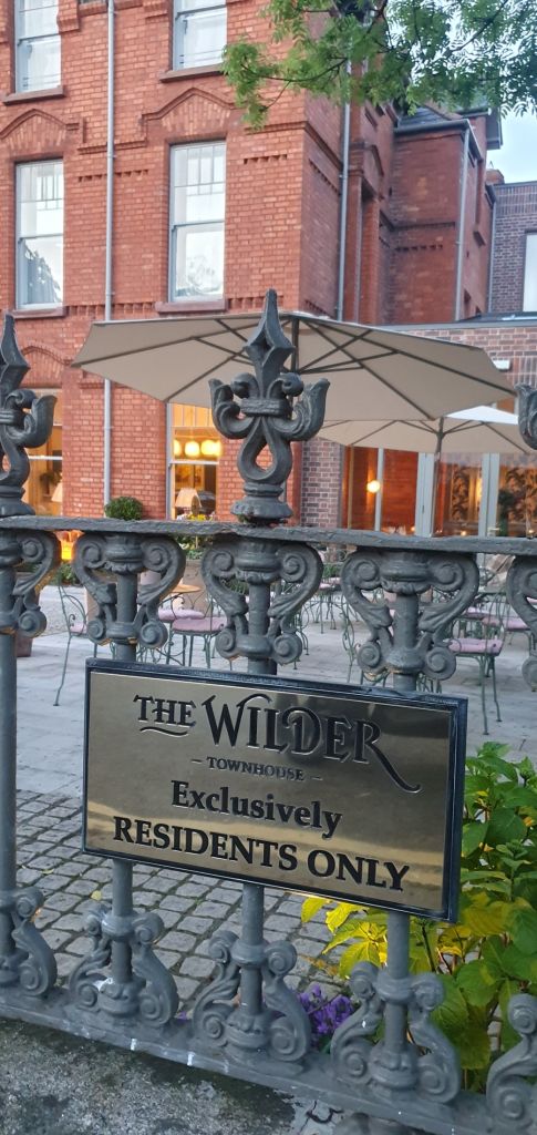 Een smeedijzeren hek met een gouden bord met de tekst The Wilder Townhouse, Exclusively Residents Only staat op de voorgrond. Achter het hek staan tafels en stoelen buiten onder grote beige parasols. Op de achtergrond maakt een bakstenen gebouw met verlichte ramen deze luxueuze herenhuissetting compleet. | Echt Ierland Vakanties