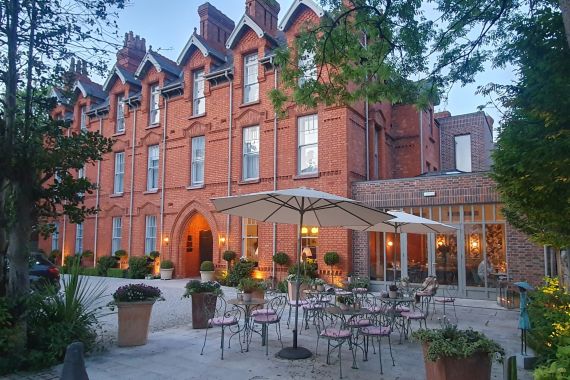 Een roodstenen hotel, het Wilder Townhouse, heeft gebogen deuren en hoge ramen. Een buitenpatio met roze stoelen, metalen tafels, grote parasols en potplanten bevindt zich op de voorgrond. Warme lichten verlichten het gebied, terwijl weelderig groen en bomen een gezellige ambiance creëren. | Echt Ierland Vakanties