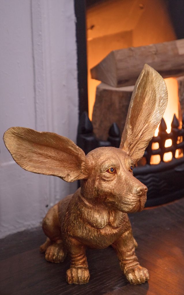 Een gouden hondensculptuur met overdreven grote oren staat op een donker oppervlak in de gezellige lounge van Wilder Townhouse. De achtergrond wordt verwarmd door een brandende open haard, knisperende houtblokken creëren een uitnodigende ambiance. De uitdrukking van de hond is kalm en attent, wat charme toevoegt aan deze elegante hotelsetting. | Echt Ierland Vakanties