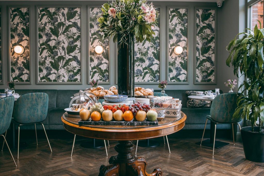 In de elegante eetkamer van Wilder Townhouse staat een ronde houten tafel met diverse soorten fruit, gebak en yoghurtbekers. In het midden staat een groot bloemstuk met roze rozen. De achtergrond bestaat uit behang met groene bladeren en donkergroene gestoffeerde stoelen rond een tafel in hotelstijl. | Echt Ierland Vakanties
