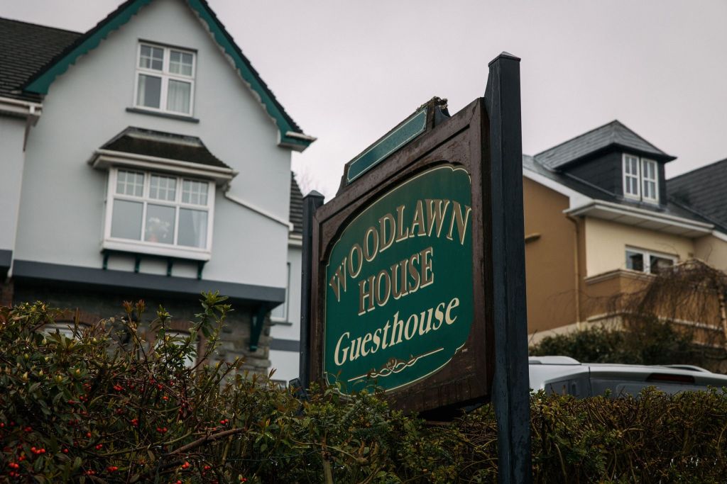 Het bord voor "Woodlawn House Guesthouse" met een donkergroen en goudkleurig ontwerp is prominent aanwezig. Erachter staat een groot huis met twee verdiepingen, witte muren en decoratieve gevels, tegen een bewolkte lucht. Omringende struiken voegen groen toe aan de scène. | Echt Ierland Vakanties