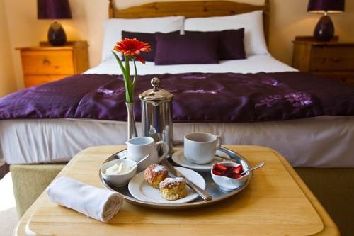 Een dienblad met thee, twee kopjes en een scone met room en jam staat op een houten tafel in Woodlawn House. Ernaast ligt een handdoek opgerold naast het dienblad. Het bed is voorzien van paars beddengoed en kussens, terwijl een enkele rode bloem in een vaas charme toevoegt. Houten nachtkastjes en paarse lampen zijn zichtbaar op de achtergrond. | Echt Ierland Vakanties