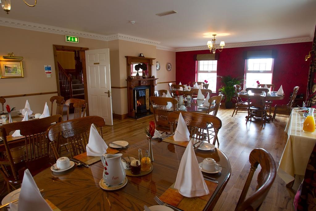 Een gezellige eetkamer die doet denken aan Woodlawn House verwelkomt u met houten vloeren en tafels die zijn gedekt voor maaltijden. Elke tafel heeft witte servetten, kopjes en schoteltjes. Een buffet met een jus d'orangekan staat rechts. De door Killarney geïnspireerde kamer heeft rode muren, een open haard, houten stoelen, grote ramen en een warme kroonluchter. | Echt Ierland Vakanties