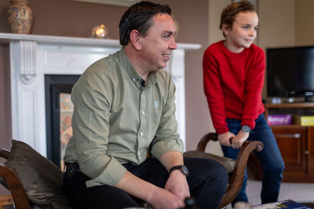Een man in een groen shirt en een jonge jongen in een rode trui zitten in een woonkamer. De man zit op een bankje, glimlachend, met zijn handen op zijn schoot. De jongen staat naast hem, licht voorovergebogen. Een open haard, tv en vaas staan op de achtergrond, wat een gezellige sfeer creëert. | Echt Ierland Vakanties