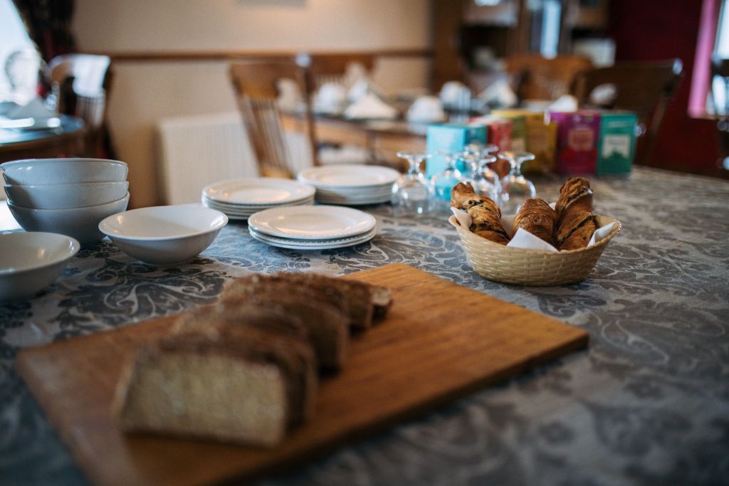 Een eettafel is gedekt met een houten plank met gesneden brood en een rieten mand met croissants. Links staan stapels witte kommen en borden. Op de achtergrond staan omgekeerde glazen en kleurrijke theedozen. De setting is gezellig, met houten stoelen en gebloemde tafelkleden. | Echt Ierland Vakanties