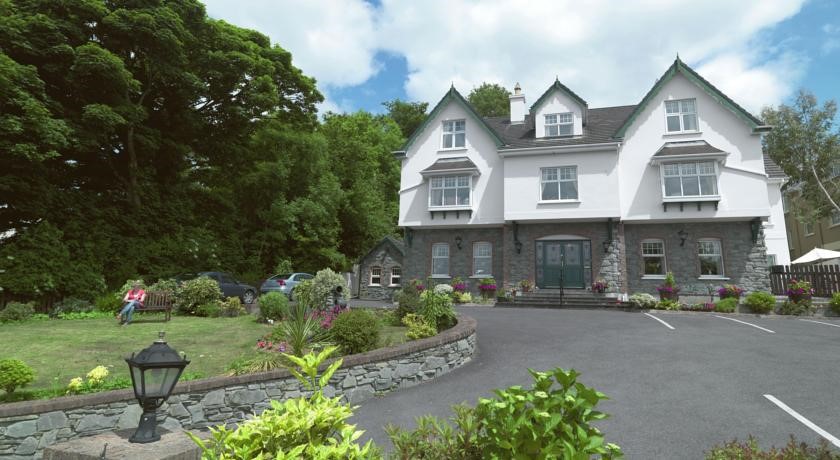 Te midden van het weelderige groen van Killarney staat Woodlawn House, een grote, witte, drie verdiepingen tellende woning met donkere dakspanen en dakkapellen. Een stenen keermuur omlijst de aangelegde tuin waar iemand op een bankje zit. De oprit, gedeeltelijk zichtbaar, herbergt geparkeerde auto's onder een gedeeltelijk bewolkte lucht. | Echt Ierland Vakanties
