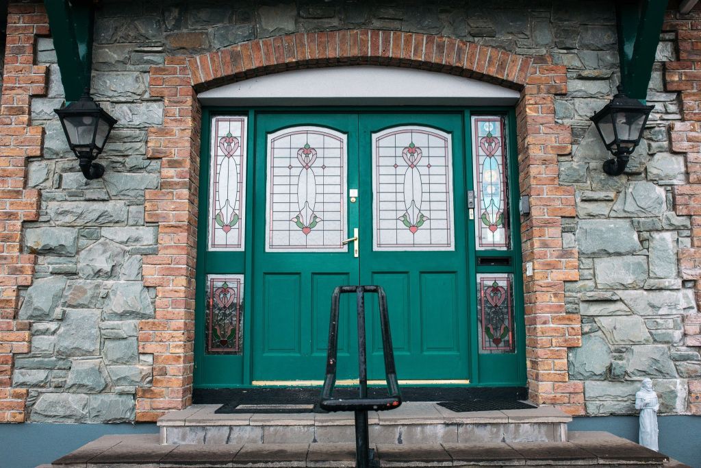 Een groene dubbele deur met ingewikkelde glas-in-loodpanelen met bloemmotieven is geplaatst in een stenen muur. Rode bakstenen omlijsten de deuropening, terwijl er aan beide kanten zwarte lantaarns zijn gemonteerd. Een kleine trede met een zwarte leuning leidt naar de ingang. | Echt Ierland Vakanties