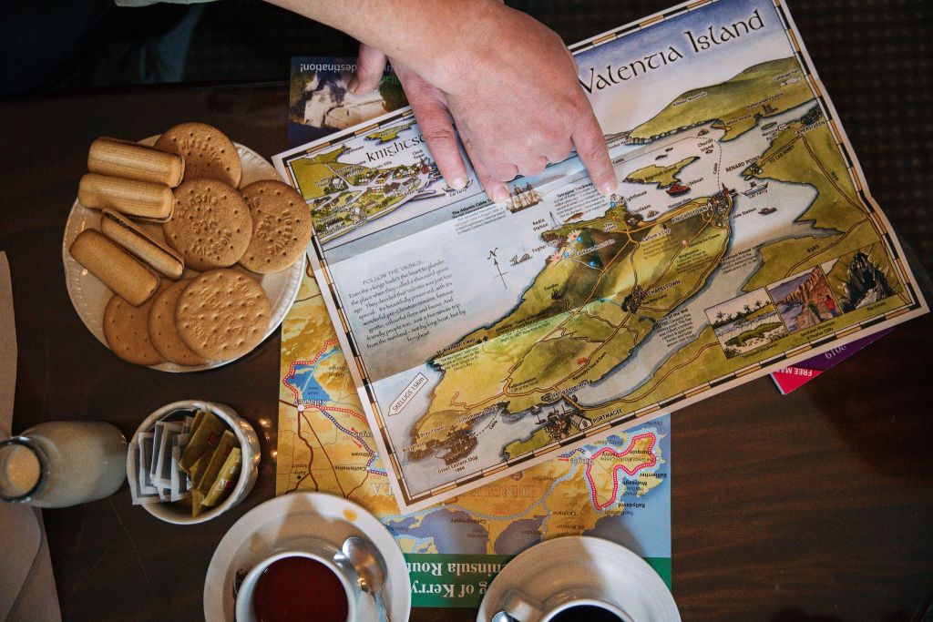 Een persoon wijst naar een kaart van Valentia Island op een tafel. In de buurt liggen een kop thee, een kop koffie, een stapel koekjes op een bord, een klein melkflesje en diverse suikerzakjes. Extra gevouwen kaarten zijn gedeeltelijk zichtbaar onder de hoofdkaart. | Echt Ierland Vakanties