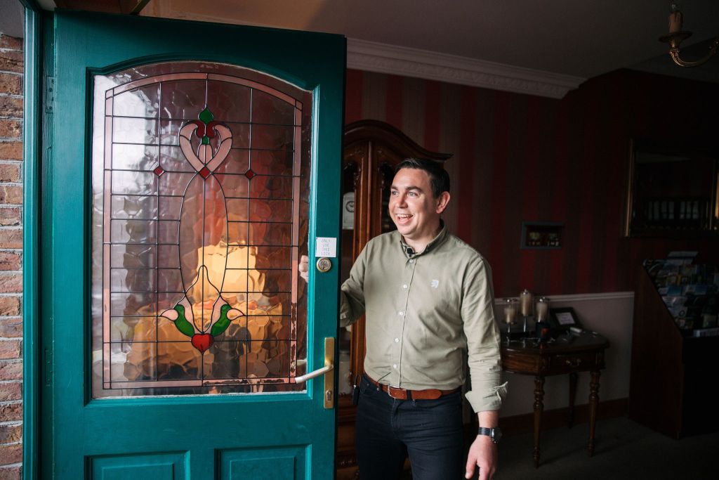 Een persoon in een groen shirt en zwarte broek staat bij de ingang en houdt de deur open. De deur heeft een glas-in-loodontwerp met hart- en bladmotieven. Het interieur heeft rood gestreept behang, een houten kast en een tafel met kaarsen. De persoon lacht. | Echt Ierland Vakanties