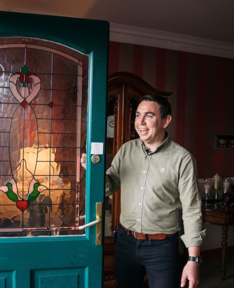 Een persoon in een groen shirt en zwarte broek staat bij de ingang en houdt de deur open. De deur heeft een glas-in-loodontwerp met hart- en bladmotieven. Het interieur heeft rood gestreept behang, een houten kast en een tafel met kaarsen. De persoon lacht. | Echt Ierland Vakanties