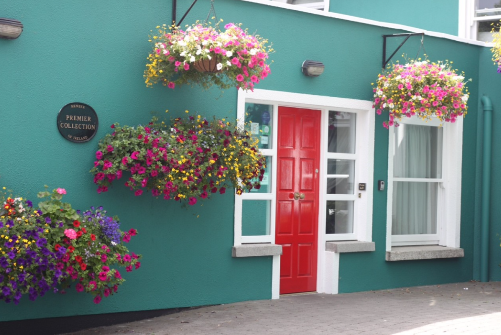 Een felrode deur in een wit frame contrasteert met een levendige groenblauwe muur. Hangende manden met kleurrijke bloemen - roze, paars, geel en wit - zorgen voor vrolijkheid bij een raam en een muurplaquette met de tekst Premier Collection. | Echt Ierland Vakanties