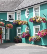 Een levendig turquoise huis met grijze dakbedekking heeft ronde en rechthoekige ramen, elk versierd met hangende manden vol kleurrijke bloemen in roze, paarse, gele en witte tinten. Links van het huis zit een rode deur. | Echt Ierland Vakanties