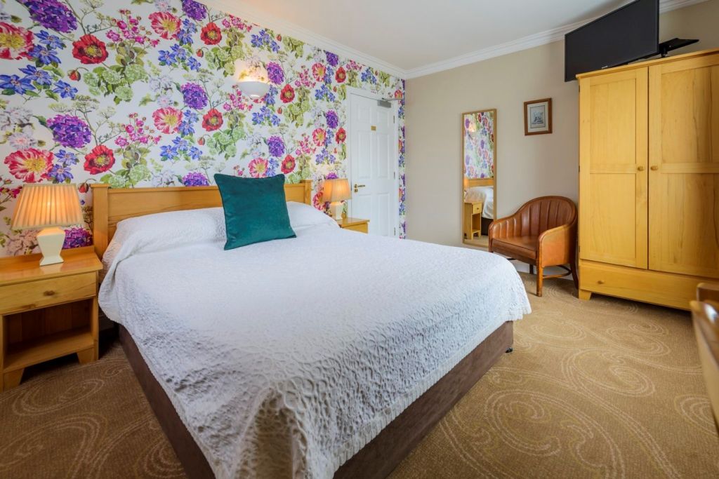 Een gezellige slaapkamer met een tweepersoonsbed bedekt met een wit dekbed, een houten hoofdeinde en twee nachtkastjes met lampen. Fel bloemenbehang siert één muur. Er is een kledingkast, een kleine spiegel, een fauteuil en een tv boven de kledingkast. | Echt Ierland Vakanties