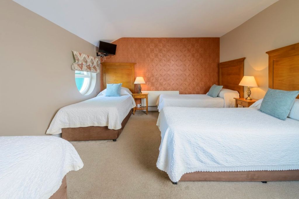 Een hotelkamer met drie eenpersoonsbedden, elk met wit beddengoed en lichtblauwe kussens. Er zijn houten hoofdeinden, twee bedlampjes, een rond raam met een kap met patroon, een kleine tv aan de muur en een accentmuur met patroon. Beige tapijt bedekt de vloer. | Echt Ierland Vakanties