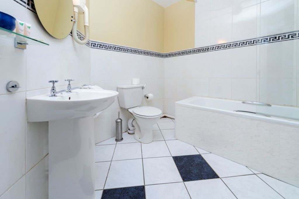 Een nette badkamer met witte en blauwe tegelvloer, wastafel op voet, toilet, toiletpapierhouder, zilveren afvalemmer en een bad met glazen scherm. De muren zijn wit met een Griekse sleutelrand en een beige bovengedeelte. Spiegel en planchet met toiletartikelen boven de wastafel. | Echt Ierland Vakanties