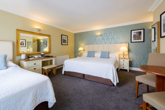 Een ruime hotelkamer met een groot tweepersoonsbed en een eenpersoonsbed, beide met witte spreien. Er zijn nachtkastjes met lampen, een spiegel boven een klein bureau met een waterkoker, ingelijste foto's aan de muren en een ronde tafel met twee stoelen. | Echt Ierland Vakanties