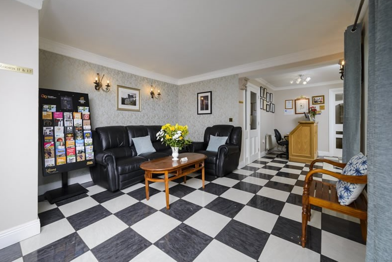 De lobby van het hotel heeft een zwart-wit geblokte vloer, zwartleren banken met lichte kussens, een houten salontafel met een vaas gele en witte bloemen, een rek met brochures, ingelijste foto's op bleke muren en een houten receptie achterin. | Echt Ierland Vakanties