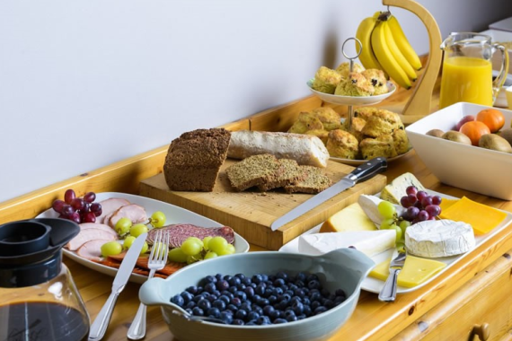 Een houten tafel gedekt met ontbijtspullen: brood, kaas, vleeswaren, druiven, scones, bananen, een schaal bosbessen, een bord met gemengd fruit, een kan sinaasappelsap, een koffiepot en bestek dat netjes was neergelegd om te serveren. | Echt Ierland Vakanties