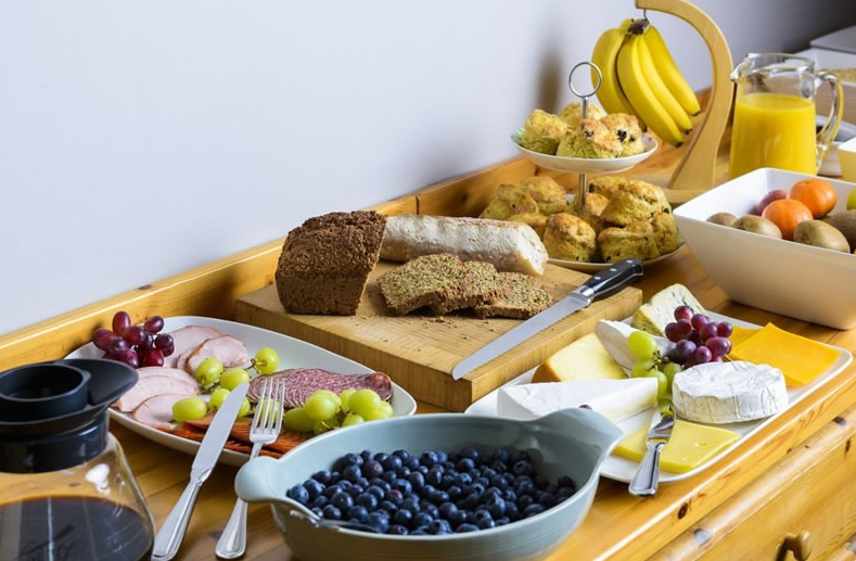 Een houten tafel gedekt met ontbijtspullen: brood, kaas, vleeswaren, druiven, scones, bananen, een schaal bosbessen, een bord met gemengd fruit, een kan sinaasappelsap, een koffiepot en bestek dat netjes was neergelegd om te serveren. | Echt Ierland Vakanties