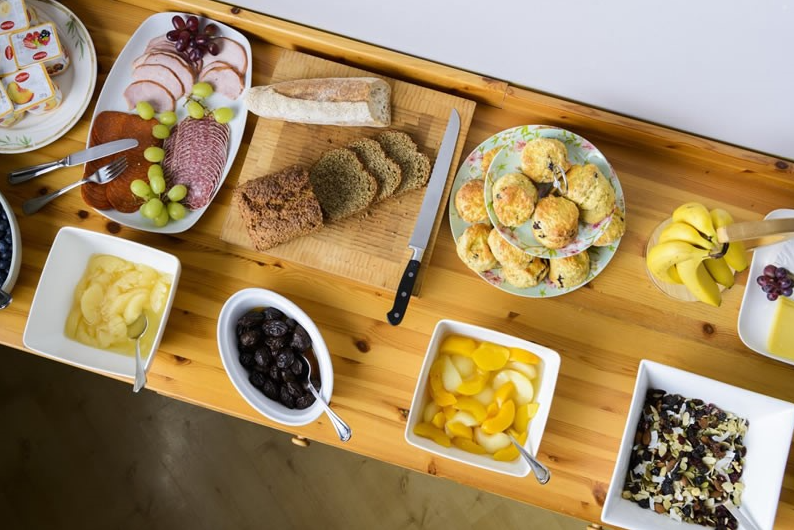 Een houten gedekte tafel met verschillende ontbijtproducten: gesneden brood, stokbrood, mes, scones op een gebloemd bord, vleeswaren met druiven, bananen, appelschijfjes, pruimen, perziken in blik, cornflakes met gedroogd fruit en boter op toast. | Echt Ierland Vakanties