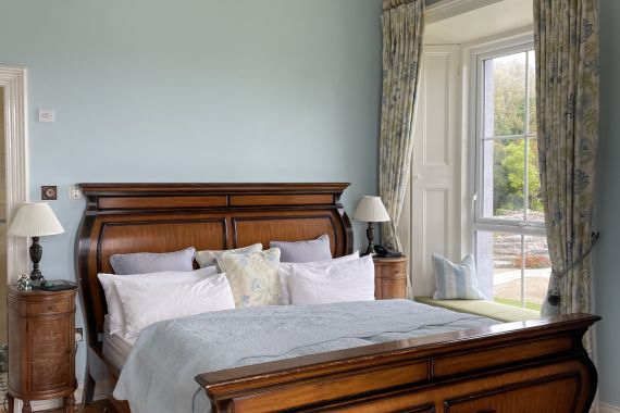 Elegante slaapkamer met een vintage houten slee bed, bedekt met wit en lichtblauw beddengoed. Een kristallen kroonluchter hangt aan het plafond. Gordijnen omlijsten een hoog raam met uitzicht op de tuin. Twee nachtkastjes met lampen flankeren het bed en een klein bagagerek staat aan het voeteneind. | Echt Ierland Vakanties