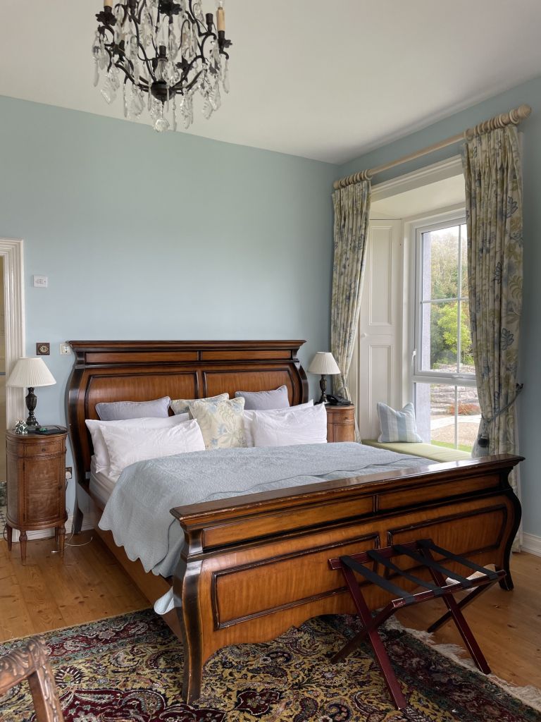 Elegante slaapkamer met een vintage houten slee bed, bedekt met wit en lichtblauw beddengoed. Een kristallen kroonluchter hangt aan het plafond. Gordijnen omlijsten een hoog raam met uitzicht op de tuin. Twee nachtkastjes met lampen flankeren het bed en een klein bagagerek staat aan het voeteneind. | Echt Ierland Vakanties