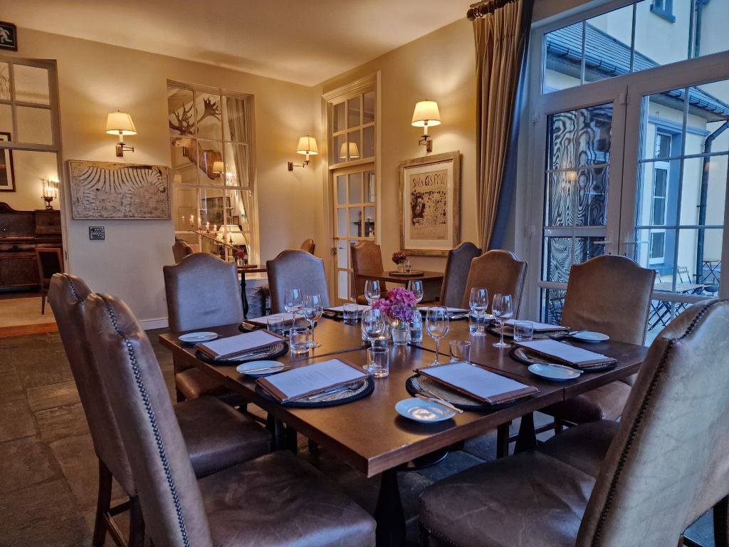Een gezellige eetkamer met een houten tafel voor acht personen, met elegante bruine leren stoelen. De tafelopstelling bestaat uit witte borden, glazen en menu's. Zachte sfeerverlichting komt van aan de muur gemonteerde lampen. Grote ramen bieden uitzicht naar buiten en kunstwerken sieren de muren. Een bloemstuk siert de tafel. | Echt Ierland Vakanties