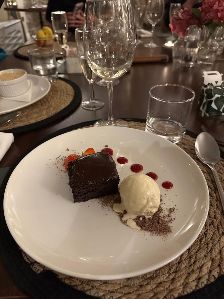 Een elegant geplateerd dessert op een ronde witte schaal bestaat uit een chocoladetaart met glanzend glazuur, een bolletje vanille-ijs en decoratieve druppels rode saus. Het wordt geserveerd op een geweven placemat met een glas witte wijn, omringd door andere eetwaren en een zachte, sfeervolle setting. | Echt Ierland Vakanties