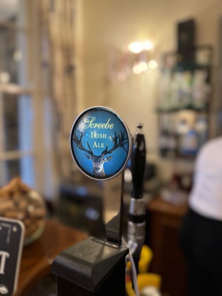 Close-up van een biertap met een rond etiket met de tekst "Screebe Irish Ale" met een silhouet van een hertenkop op een blauwe achtergrond. De tap staat in een baropstelling met planken vol flessen op de wazige achtergrond en een persoon gedeeltelijk zichtbaar aan de rechterkant. | Echt Ierland Vakanties