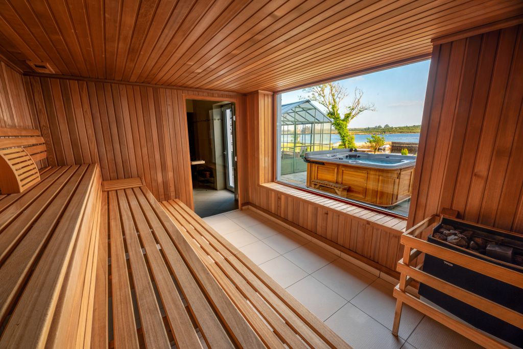 Ruim houten sauna-interieur met een lange bank aan de linkerkant en een kachel aan de rechterkant. Een groot raam toont een uitzicht naar buiten op een glazen kas en een houten hottub, met een schilderachtig meer en bomen op de achtergrond onder een heldere hemel. Zonlicht vult de gezellige ruimte. | Echt Ierland Vakanties