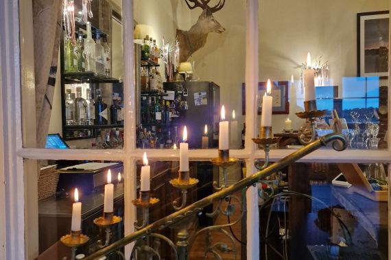 Kijk door een raam naar een warm verlichte kamer met een kandelaar met brandende kaarsen. De kamer heeft een bar vol flessen, een gemonteerde hertenkop, ingelijste kunst aan de muur en een wijnrek met flessen. Reflecties zijn te zien op het glas, wat bijdraagt aan de gezellige sfeer. | Echt Ierland Vakanties
