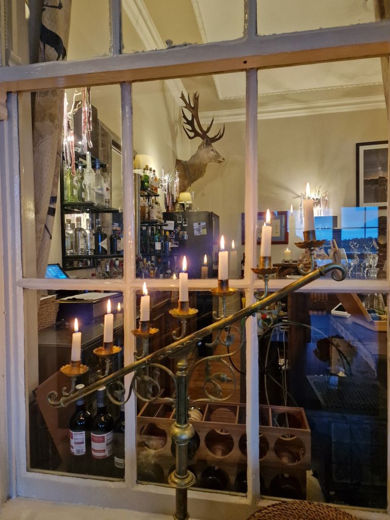 Kijk door een raam naar een warm verlichte kamer met een kandelaar met brandende kaarsen. De kamer heeft een bar vol flessen, een gemonteerde hertenkop, ingelijste kunst aan de muur en een wijnrek met flessen. Reflecties zijn te zien op het glas, wat bijdraagt aan de gezellige sfeer. | Echt Ierland Vakanties