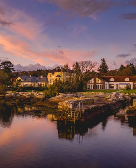 Schilderachtig uitzicht op een serene oever van het meer bij zonsondergang, met charmante huizen met grote ramen en stenen treden die naar het water leiden. Bomen en wolken weerspiegelen in het kalme oppervlak, terwijl de lucht is geschilderd met tinten roze, paars en oranje, wat een rustige sfeer creëert. | Echt Ierland Vakanties