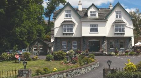 Het twee verdiepingen tellende Woodlawn House heeft groene afwerking, witte muren en stenen accenten die doen denken aan de charme van Killarney. Een lage stenen muur grenst aan de oprit die naar een groene deur leidt. De weelderige tuin toont kleurrijke bloemen en een zittende figuur aan de linkerkant, omlijst door bomen onder een bewolkte lucht. | Echt Ierland Vakanties