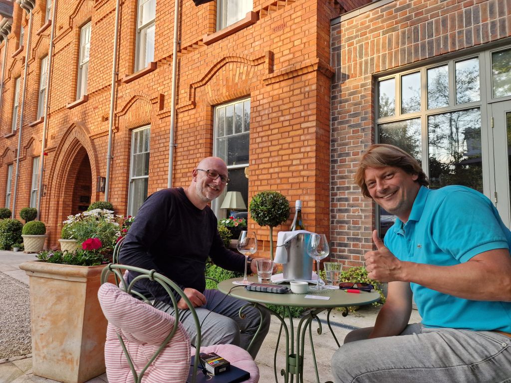 Twee mannen zitten lachend aan een klein tafeltje buiten. De een draagt een zwart shirt en een bril, de ander een turquoise poloshirt. Ze hebben wijnglazen en een wijnkoeler op tafel staan. Het geheel bevindt zich voor een rood bakstenen gebouw met boogramen. Potplanten en stoelen met kussens zorgen voor een gezellige sfeer. | Echt Ierland Vakanties
