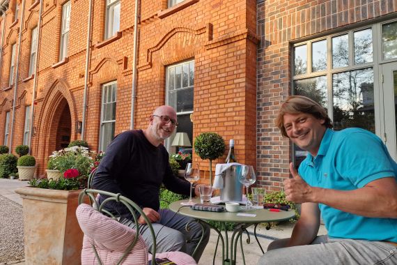 Twee mannen zitten lachend aan een klein tafeltje buiten. De een draagt een zwart shirt en een bril, de ander een turquoise poloshirt. Ze hebben wijnglazen en een wijnkoeler op tafel staan. Het geheel bevindt zich voor een rood bakstenen gebouw met boogramen. Potplanten en stoelen met kussens zorgen voor een gezellige sfeer. | Echt Ierland Vakanties