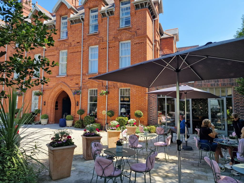 Een bakstenen gebouw met een boogvormige ingang vormt de achtergrond voor een terras. Verschillende tafels met roze stoelen met kussens staan in de schaduw van grote parasols. Potplanten en hangende bloemen sieren het terras en zorgen voor een ontspannen sfeer. Mensen zitten te genieten van de omgeving. | Echt Ierland Vakanties