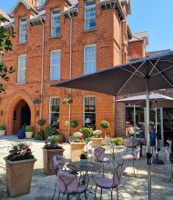Een bakstenen gebouw met een boogvormige ingang vormt de achtergrond voor een terras. Verschillende tafels met roze stoelen met kussens staan in de schaduw van grote parasols. Potplanten en hangende bloemen sieren het terras en zorgen voor een ontspannen sfeer. Mensen zitten te genieten van de omgeving. | Echt Ierland Vakanties
