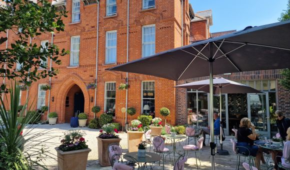 Een bakstenen gebouw met een boogvormige ingang vormt de achtergrond voor een terras. Verschillende tafels met roze stoelen met kussens staan in de schaduw van grote parasols. Potplanten en hangende bloemen sieren het terras en zorgen voor een ontspannen sfeer. Mensen zitten te genieten van de omgeving. | Echt Ierland Vakanties