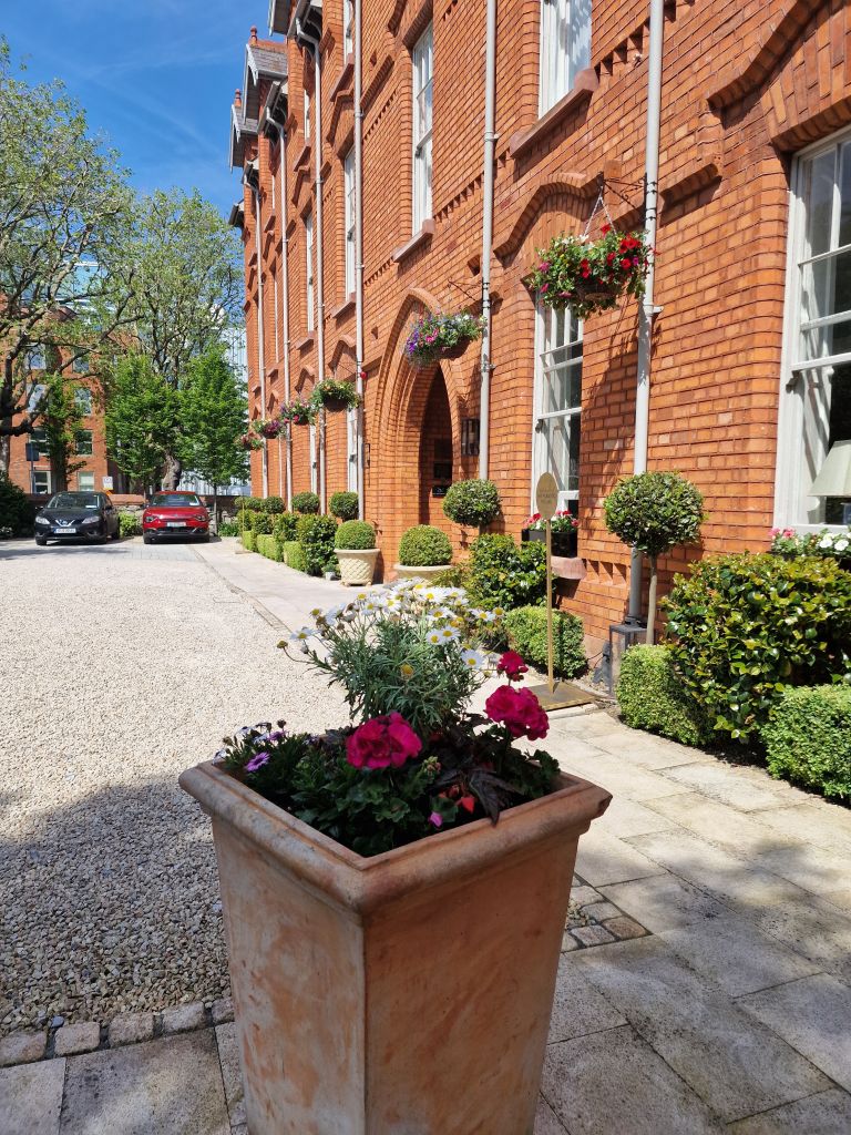 Op de voorgrond staat een terracotta plantenbak met roze, witte en paarse bloemen. Daarachter leidt een grindpad naar een roodstenen gebouw met boogramen en hangende bloemmanden. Bomen met groene bladeren staan langs de zijkant en twee geparkeerde auto's zijn zichtbaar onder een strakblauwe hemel. | Echt Ierland Vakanties
