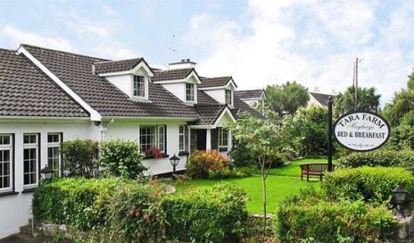 Charmant wit huis met een donker pannendak, dakkapellen en weelderige groene tuinen. Een houten bank en gesnoeide heggen sieren de voortuin. Een bord met de tekst "Tara Farm Bed & Breakfast" staat te midden van volgroeide bomen en kleurrijke bloemen onder een gedeeltelijk bewolkte hemel. | Echt Ierland Vakanties