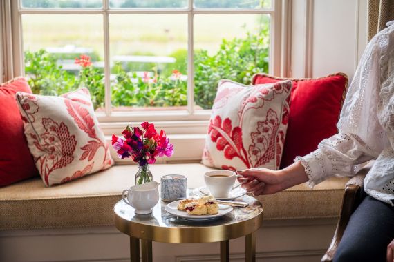 Een knusse vensterbank met rode en witte bloemenkussens, een klein rond tafeltje met een theekopje, een scone met jam en room, een wit kopje, een mok met print en een vaasje met roze bloemen. Iemands hand houdt het theekopje vast. Buiten het raam is groen te zien. | Echt Ierland Vakanties