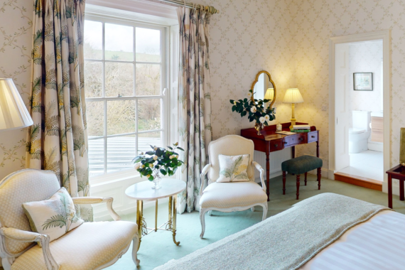 Een lichte, elegante slaapkamer met bloemenbehang, twee witte fauteuils en een kleine ronde tafel bij het raam. Een bed met een groene sprei staat tegenover een houten kaptafel met een spiegel en een lamp. Een open deur leidt naar een zonnige aangrenzende kamer met meer meubels. | Echt Ierland Vakanties