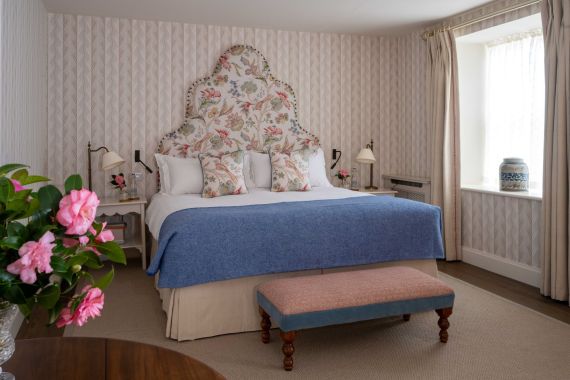Een knusse slaapkamer met een hoofdbord met bloemenmotief, bijpassende kussens, een blauwe deken op het bed en een roze gestoffeerde bank aan het voeteneind. Een vaas met roze bloemen staat op een houten tafel en zonlicht filtert door een raam met gordijnen aan de rechterkant. | Echt Ierland Vakanties